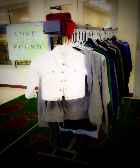 Small white child's jacket hung on a lost and found rack at school with other larger clothing items.